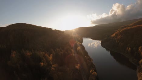 Cinematic-aerial-diving-shot-of-a-straight-road-following-a-scenic-water-dam-surrounded-by-lush-autumn-trees