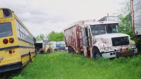 Weite-Aufnahme-Eines-Alten-Schulbusses-Und-Eines-Müllwagens,-Die-Im-Gras-Stehen-Und-Den-Elementen-Ausgesetzt-Sind