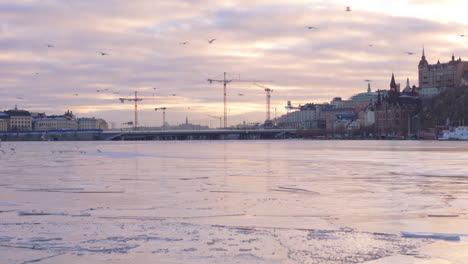 Rückzug-Der-Drohne-In-Der-Dämmerung-über-Dem-Gefrorenen-Riddarfjärden,-Blick-Auf-Kräne-über-Der-Schleusenbrücke