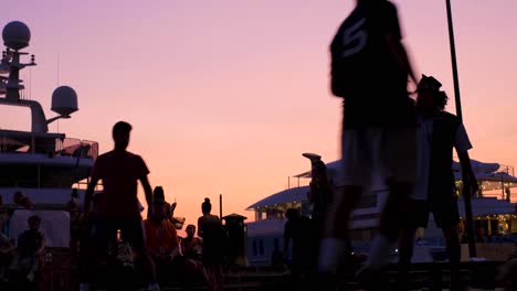 Vista-Cercana-En-Cámara-Lenta-De-Las-Siluetas-De-Los-Jugadores-Durante-El-Torneo-De-Fútbol-Callejero-Al-Atardecer-Con-Yates---Espectadores-En-Segundo-Plano