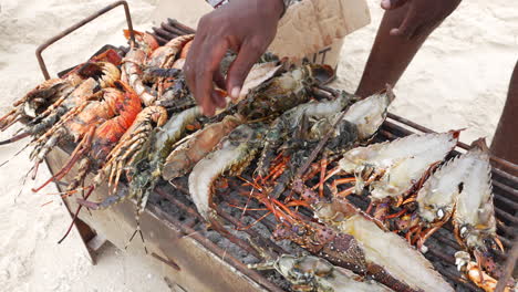 Langostas-Y-Langostinos-Cocinados-En-Una-Pequeña-Parrilla-En-La-Playa,-Un-Hombre-Local-Pesca-Con-La-Mano,-Zanzíbar,-Tanzania,-Filmado-A-30-Fps