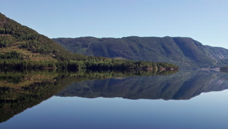 Panorámica-Aérea-Sobre-Las-Tranquilas-Aguas-De-Byglandsfjord-En-Noruega-En-Una-Mañana-Soleada,-El-Cielo-Azul-Y-Las-Montañas-Cubiertas-De-Bosques-Se-Reflejan-En-El-Agua-Que-Ondula-Suavemente