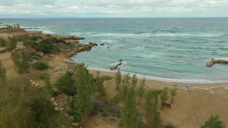 Overhead-shot-of-a-coastal-village-with-white-houses,-blue-beach-umbrellas,-and-a-curving-sandy-shoreline,-adjacent-to-rocky-outcrops-and-turbulent-sea-waters