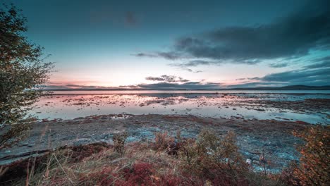 Stormy-dark-clouds-move-fast-in-the-sunset-sky-above-the-rocky-coast-with-exposed-sandbank-covered-with-weeds-and-kelp