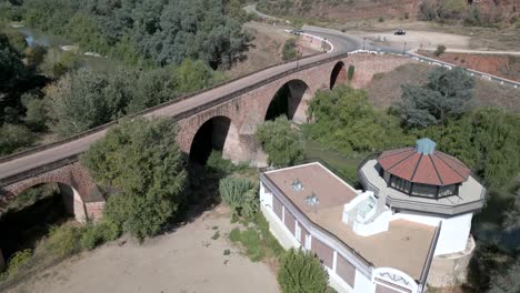 Un-Dron-Captura-El-Edificio-Del-Spa-Y-Sus-Exuberantes-Jardines,-Con-El-Telón-De-Fondo-De-Un-Puente-Del-Siglo-XVI-Que-Cruza-El-Río-Guadalquivir,-Todo-Parte-Del-Centro-De-Spa-Marmolejo,-Andalucía,-España