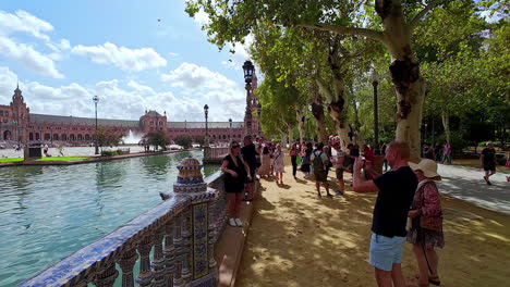 Los-Turistas-Caminan-Por-La-Plaza-De-España,-Sevilla,-España,-Un-Lugar-Emblemático-De-Andalucía,-Destino-De-Viaje,-Tomando-Fotografías-De-La-Arquitectura-Colonial-Junto-Al-Río-Limpio.