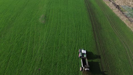 Aerial-Drone-Shot-of-Tractor-Spraying-Manure-on-Green-Grass-Field-with-Snowy-Mountains-in-Background