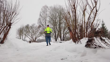 Male-in-green-safety-jacket-walk-on-snowy-countryside-path-with-bare-tree-aside