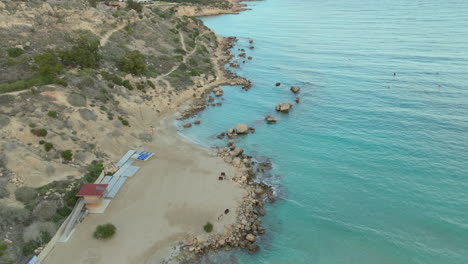 La-Imagen-Aérea-Captura-Un-Suave-Arco-De-Playa-De-Arena-Escondido-Entre-Escarpados-Acantilados-Y-Las-Aguas-Azules-Del-Mediterráneo,-Con-Un-Puñado-De-Rocas-A-Lo-Largo-De-La-Costa-E-Instalaciones-De-Playa-Cercanas.