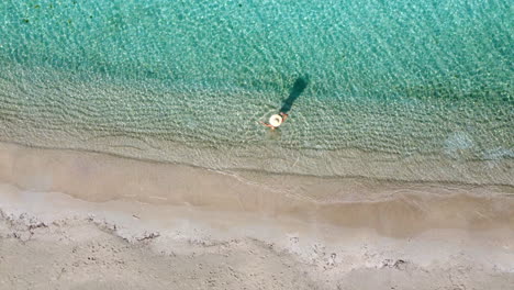 Aerial-top-down-view-of-a-woman-with-straw-hat-in-turquoise-sea-next-to-a-tropical-empty-paradise-beach
