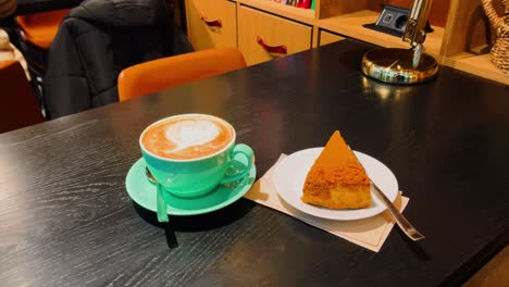 Primer-Plano-De-Una-Taza-De-Café-Con-Leche-Y-Un-Trozo-De-Tarta-De-Queso-Con-Caramelo-Sobre-Una-Mesa-Negra-En-La-Cafetería