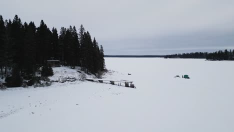 A-Slow-Drone-Landscape-Shot-of-an-Island-on-Frozen-Canadian-Paint-Lake-with-an-Ice-fishing-hut-and-skioos