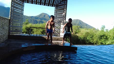 Two-kids-jumping-into-the-swimming-pool-surrounded-with-bamboo-trees-and-mountainous-background,-slow-motion