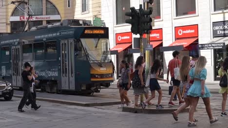 Una-Multitud-De-Personas-Camina-Por-Un-Cruce-En-La-Ciudad-De-Milán,-Con-Un-Tranvía-Al-Fondo