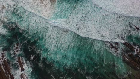 overhead-aerial-shot-over-the-Cantabrian-coast-with-linear-geological-remains-in-maroon-color-where-the-relaxing-waves-of-turquoise-colors-with-white-foam-break-in-Cantabria-Spain