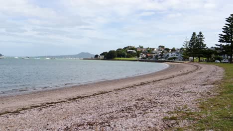 A-handheld-shot-of-a-calm-beach-with-small-waves-and-a-group-of-boats,-with-a-volcano-in-the-background,-on-a-windy-and-cloudy-day-in-a-quiet-neighborhood-in-Auckland,-New-Zealand