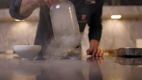 Chef-lifts-glass-cloche-to-reveal-food-with-swirling-smoke-or-steam,-gourmet-kitchen