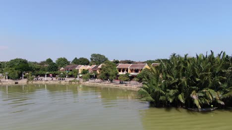 Drone-aerial-view-in-Vietnam-flying-over-Hoi-An-brown-color-river-canal-in-the-city-in-front-of-small-houses-and-palm-trees-on-a-sunny-day