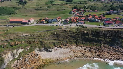 aerial-shot-on-the-coast-of-the-Cantabrian-Sea-towards-the-cliff-there-is-a-road-on-which-cars-pass-next-to-typical-houses-in-green-meadows-with-the-sky-in-the-background-and-clouds-in-Cantabria-Spain