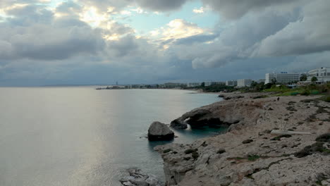 Ajia-Napa---Overcast-coastal-view-with-rocky-shoreline,-calm-sea,-and-a-cityscape-in-the-distance-under-a-dramatic-sky---aerial