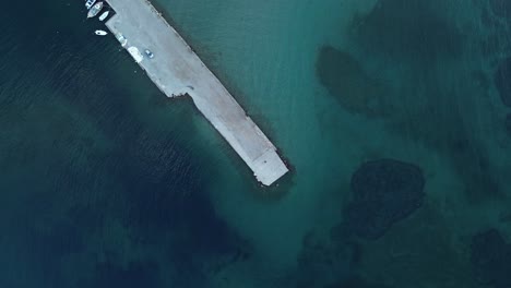 Drone-shot-straight-down,-capturing-a-coastal-scene-with-a-concrete-pier,-some-boats,-and-turquoise-seawater,-all-along-a-beach-in-Greece