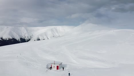 Snow-covered-Gainatu-Refuge-with-Papusa-Peak-in-the-clouds,-Iezer-Papusa-Mtns,-Arges,-Romania,-hikers-in-white-landscape