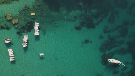 Top-down-aerial-overview-pans-across-small-skiff-and-sailboats-anchored-below-rocky-cliffs-of-Corfu-Greece