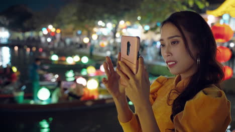 Woman-in-yellow-Ao-Dai-taking-photos-at-night-in-vibrant-Hoi-An-town,-illuminated-lanterns-on-boats