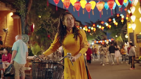 Woman-in-yellow-Ao-Dai-walking-with-bicycle-in-Hoi-An,-colorful-lanterns-at-dusk