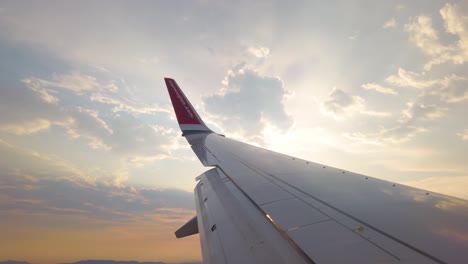 Wing-of-turning-Norwegian-airplane,-cloudy-sunset-over-city-and-hills