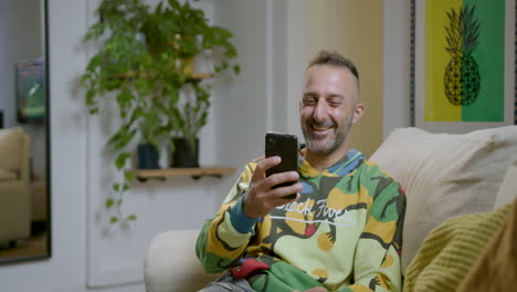 Caucasian-Man-Sitting-On-Couch,-Laughs-While-Watching-Funny-Video-On-Mobile-Phone