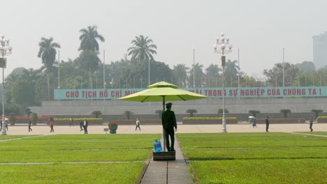 Guardia-Bajo-Un-Paraguas-Verde-Junto-Al-Mausoleo-De-Ho-Chi-Minh-En-Hanoi,-Durante-El-Día-Con-Cielos-Despejados,-Plano-Amplio