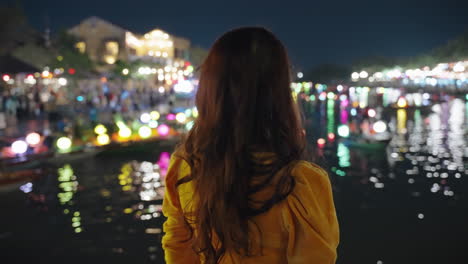 Woman-in-yellow-Ao-Dai-admires-colorful-lanterns-on-Hoi-An-river-at-night,-vibrant-cultural-scene