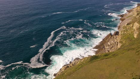 flight-with-a-drone-on-a-cliff-in-the-Cantabrian-Sea-leaving-a-green-meadow-watching-the-waves-crash-on-the-rocks-with-a-variety-of-blue-colors-in-the-sea-on-a-summer-afternoon-in-Cantabria-Spain