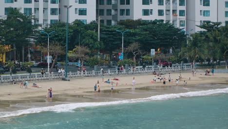 People-Walking-at-Hon-Chong-Public-Beach-at-Sunset-in-Nha-Trang-City,-vehicles-traffic-on-the-street-next-to-apartment-buildings-Vietnam---distant-view-slow-motion