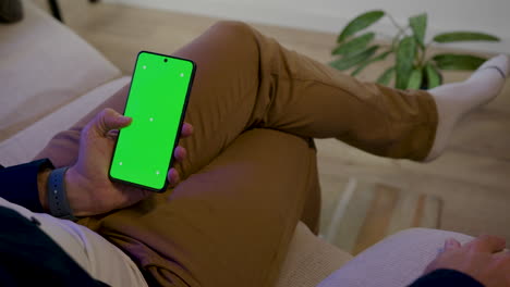 Detalle-De-Una-Mano-Masculina-Usando-Un-Teléfono-Celular-Sentado-En-El-Sofá-Mientras-Se-Relaja---Pantalla-Del-Teléfono-Celular-En-Clave-De-Croma-Verde-Para-Inserción-Gráfica