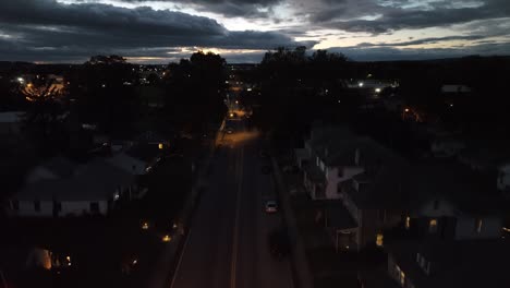 Car-driving-towards-rising-drone-on-town-street-at-night