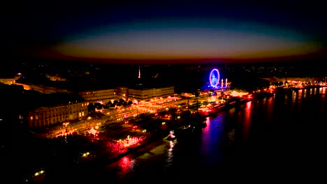 Neon-illuminated-Ferris-wheel-and-Garonne-river-shore-at-dusk-in-Bordeaux-France-during-Wine-Fair,-Aerial-pan-left-shot