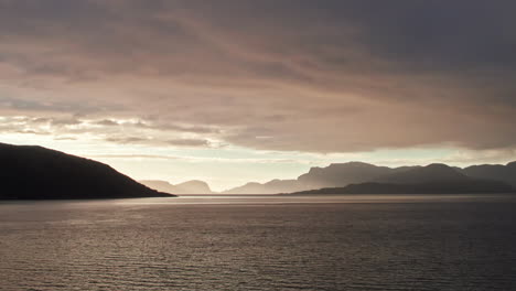Panoramic-aerial-shot,-sweeping-over-Sognefjord-in-Norway-towards-a-bright-sunset-over-the-mountainous-horizon