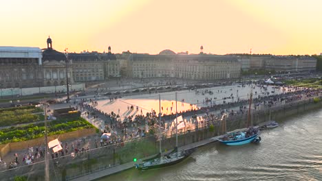 Gente-Reunida-En-La-Plaza-Place-De-La-Bourse-Durante-La-Feria-Del-Vino-Con-Veleros-En-El-Muelle,-Tiro-Aéreo-A-La-Derecha