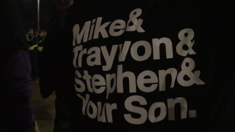 A-person-wears-a-black-T-shirt-that-reads-whit-writing,-“Mike-and-Trayvon-and-Stephen-and-your-son”-on-a-nighttime-protest