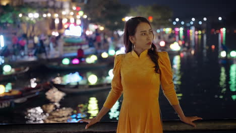 Woman-in-vibrant-Ao-Dai-posing-by-Hoi-An's-lantern-lit-river-at-night,-reflection,-cultural-attire