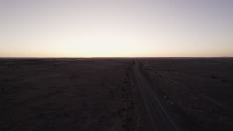 A-car-driving-down-an-empty-desert-highway-at-sunset,-drone-aerial
