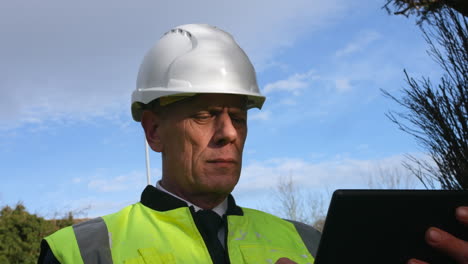 Close-up-of-a-senior-architect-examining-plans-of-a-large-building-on-a-tablet-on-a-construction-site-in-a-residential-street-with-traffic-on-the-road-in-the-background