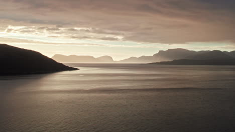 Aerial-shot,-climbing-over-the-gently-rippling-water-of-Sognefjord-in-Norway,-looking-out-at-a-sunset-over-multiple-shadowy-mountains