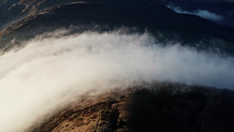 Dense-clouds-rolling-over-mountainous-terrain-at-dusk,-aerial-view