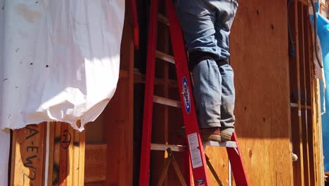 Gimbal-booming-up-shot-of-a-construction-worker-on-a-ladder-using-a-hole-cutting-drill-at-a-modular-housing-development-site-in-West-Los-Angeles,-California