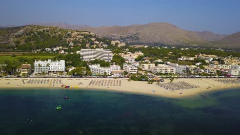 Dron-Panorámico-De-Derecha-A-Izquierda-Del-Encuadre,-Que-Muestra-El-Complejo-Playero-De-Playa-Del-Port-Pollença,-En-Mallorca,-España,-Con-Las-Montañas-Y-Los-Hoteles-De-Lujo-Al-Fondo.