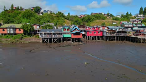 Colorful-stilt-houses-on-a-muddy-shore-during-low-tide-in-Castro,-Chiloé,-with-lush-greenery-in-the-background