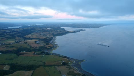 Drone-overview-of-rural-fields-and-a-Aquaculture-farm-on-the-coastline-of-Chiloe,-gloomy-evening